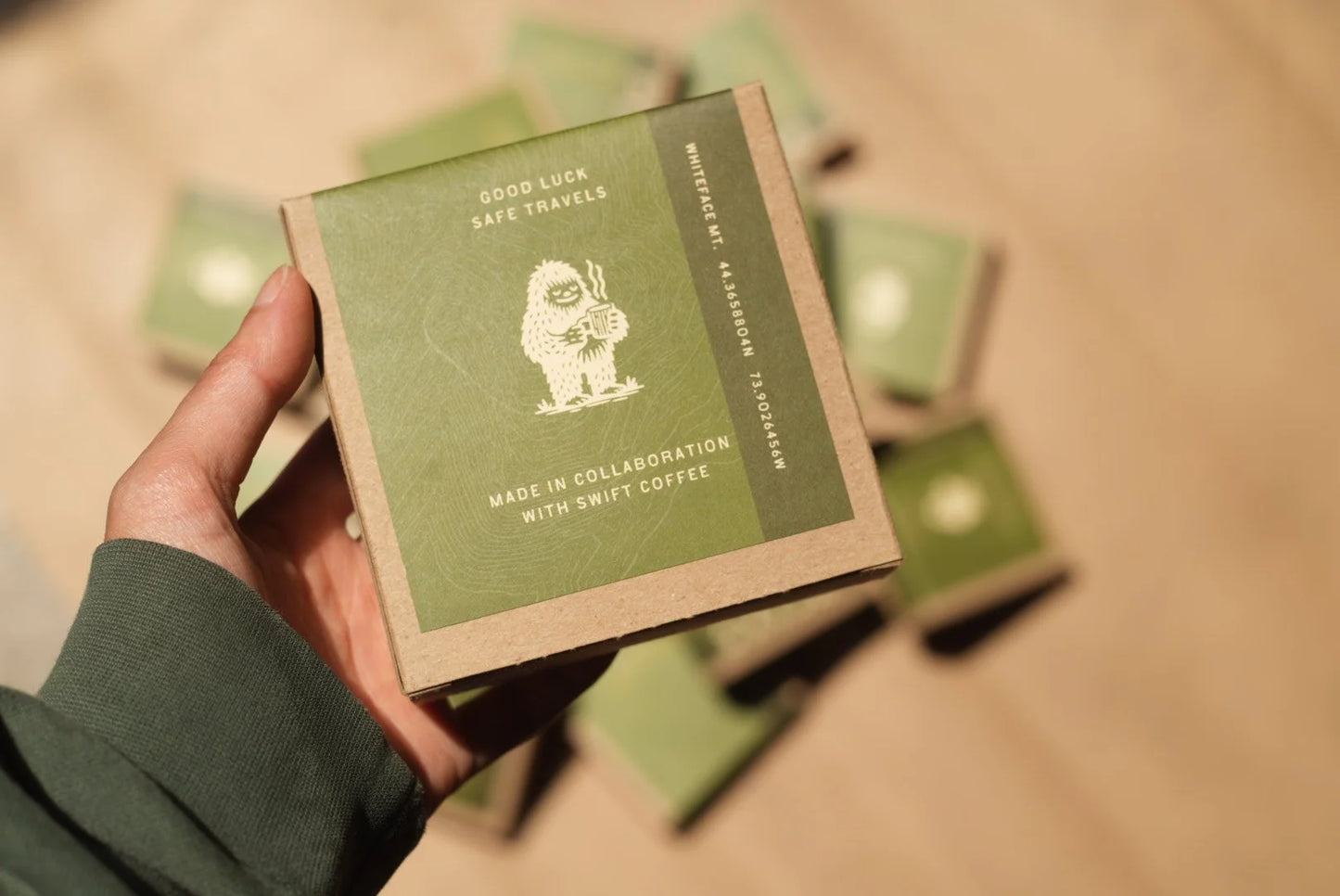 Hand holding a green and brown box with branding on a beige surface