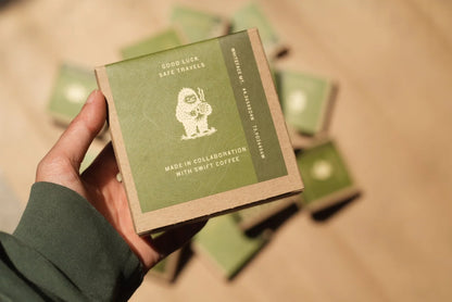 Hand holding a green and brown box with branding on a beige surface