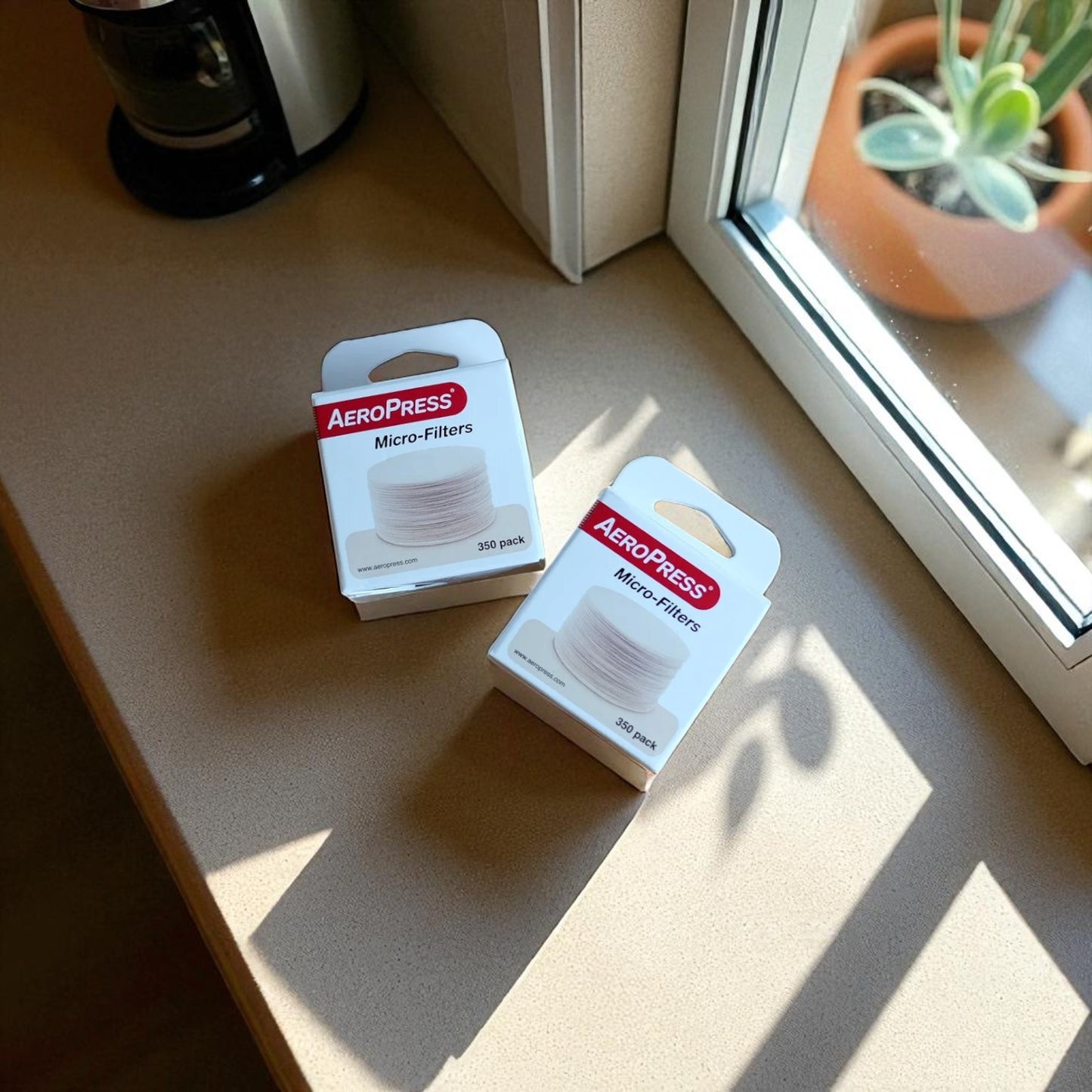 Two boxes of Aeropress micro-filters on a wooden surface with a plant and coffee maker in the background.